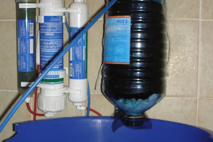 Reverse-osmosis water filter mounted on a wall, filling a blue storage container