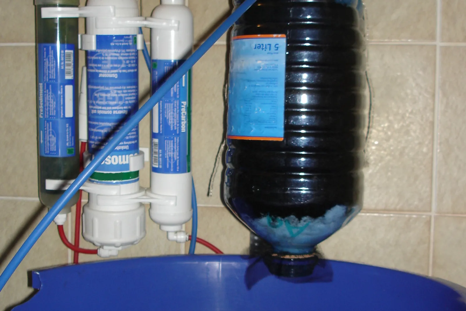 Reverse-osmosis water filter mounted on a wall, filling a blue storage container